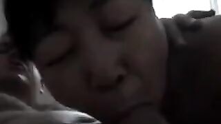 An adult Chinese woman practices her blowjob skills on him in a steamy bedroom scene